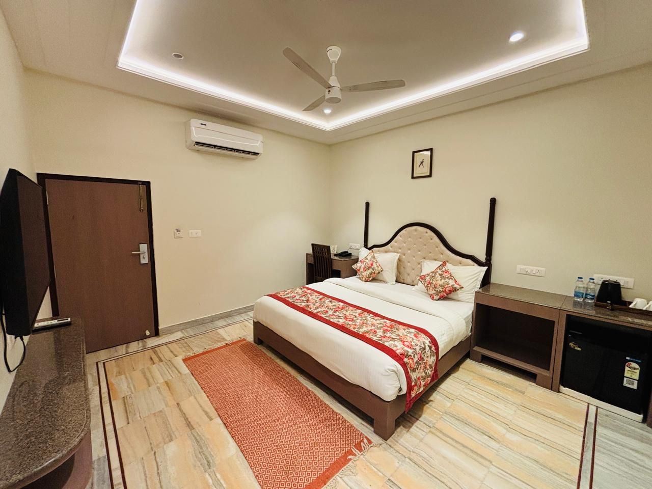 A hotel room with a bed, bedside tables, a desk, a television, an air conditioning unit, and a ceiling fan.