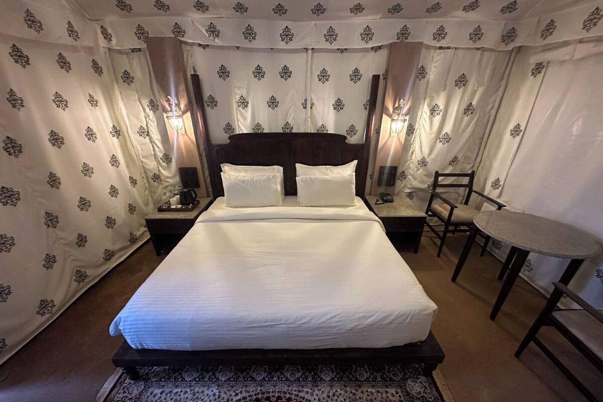 Una cama bien hecha con sábanas blancas, un cabecero de madera y una alfombra estampada. Hay una mesita con una silla, una mesita de noche con una lámpara y un teléfono instalado en la pared.