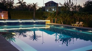 Una piscina con bordo piastrellato e cascata, circondata da palme e una casa sullo sfondo.