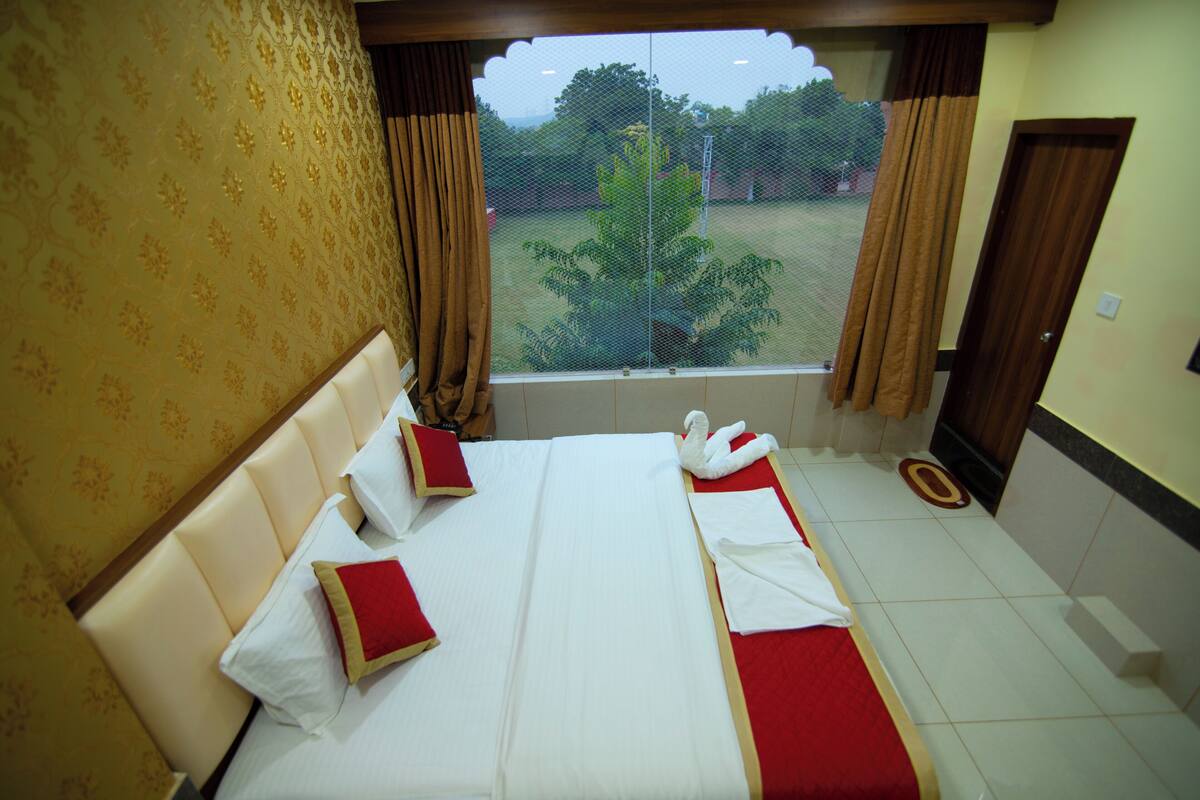 Habitación de hotel con una cama grande, dos almohadas, una manta roja y vista a un jardín por la ventana.