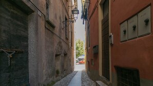 A narrow cobblestone alley between buildings with a metal gate on the left and a red building on the right. - A Due Passi dal Lago (Tremezzina)