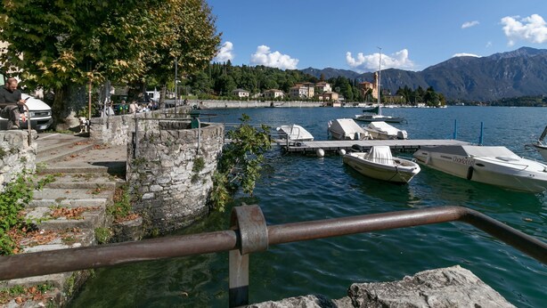 A lakeside dock with boats, stone steps, and a view of buildings and mountains in the background. - A Due Passi dal Lago (Tremezzina)