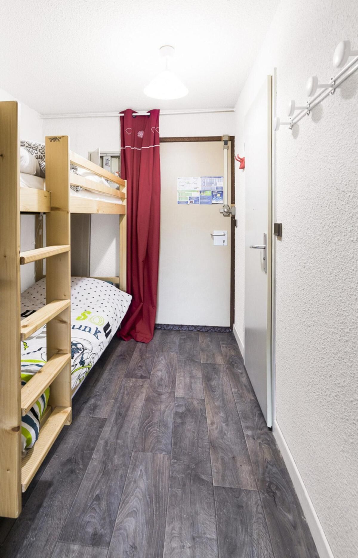 A narrow hallway with a wooden bunk bed on the left, a red curtain, and a door with a bulletin board on the right.