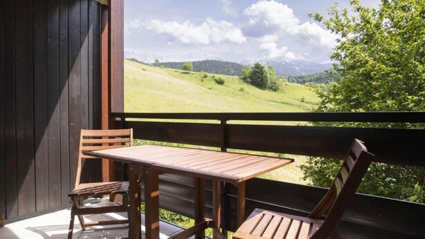 A wooden balcony with a table and chairs, overlooking a green landscape with mountains in the distance.