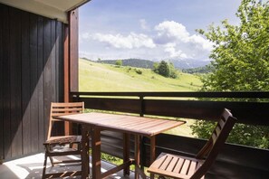 Un balcon en bois, agrémenté d’une table et de chaises, qui offre une vue sur un paysage verdoyant avec des montagnes à l’horizon.