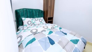 Una cama con cabecera verde, almohada estampada y cubrecama geométrico. Se observa un armario de madera adosado a la pared.