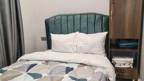 Una cama bien hecha con una colcha estampada, un cabecero tapizado de color verde y un armario de madera con ropa doblada.