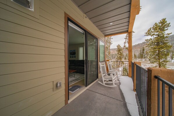 A covered balcony with a sliding glass door leading to an interior room, a wooden railing, and a snowy landscape in the background. - Modern Mountain Stay | Lake + Winter Fun (Keystone)
