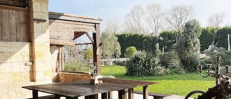 Un patio al aire libre con mesas y bancos de madera, un muro de piedra y un jardín con árboles y arbustos.