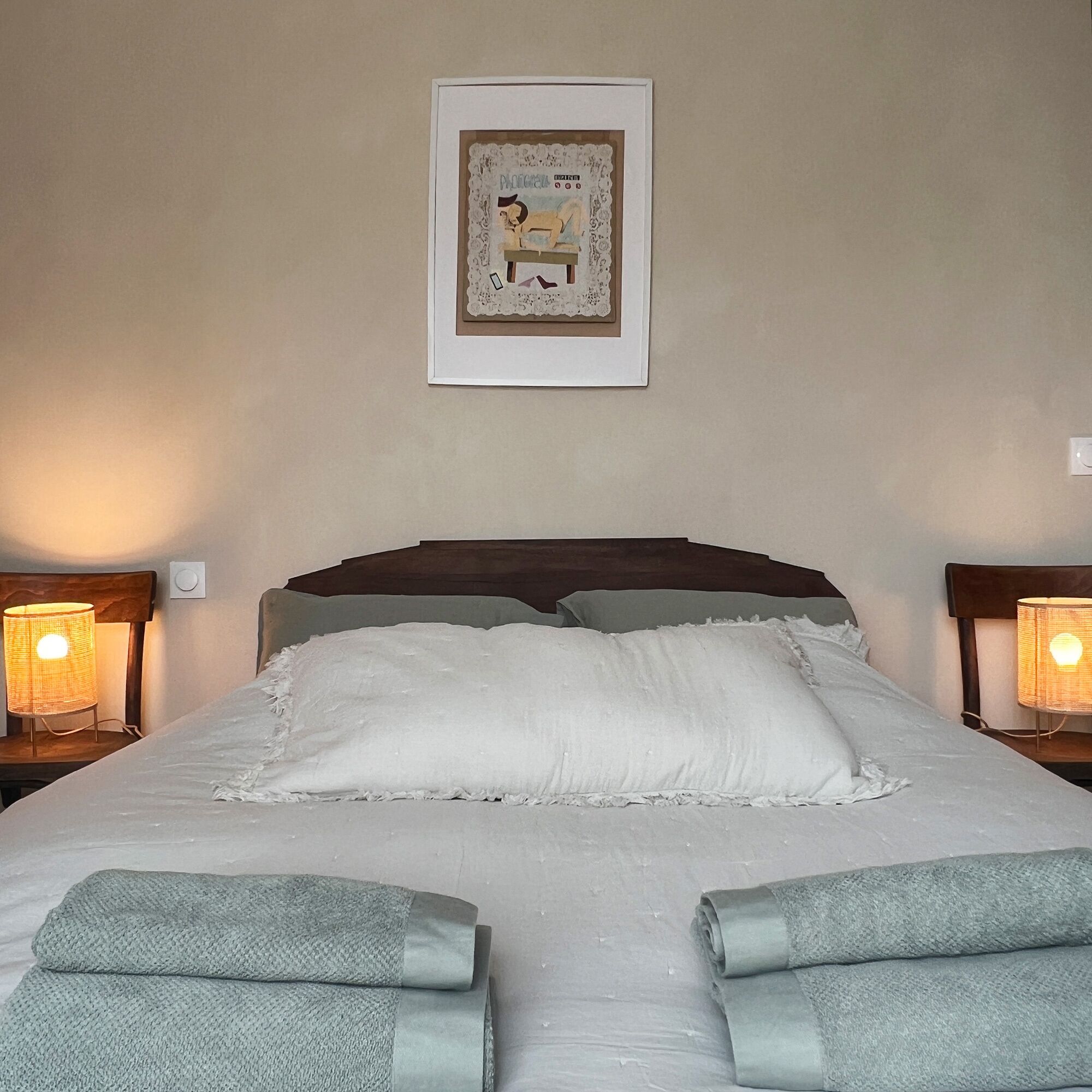 Una cama bien hecha con sábanas blancas y dos almohadas, con dos lámparas de noche a cada lado. Un cuadro enmarco cuelga sobre la cama.