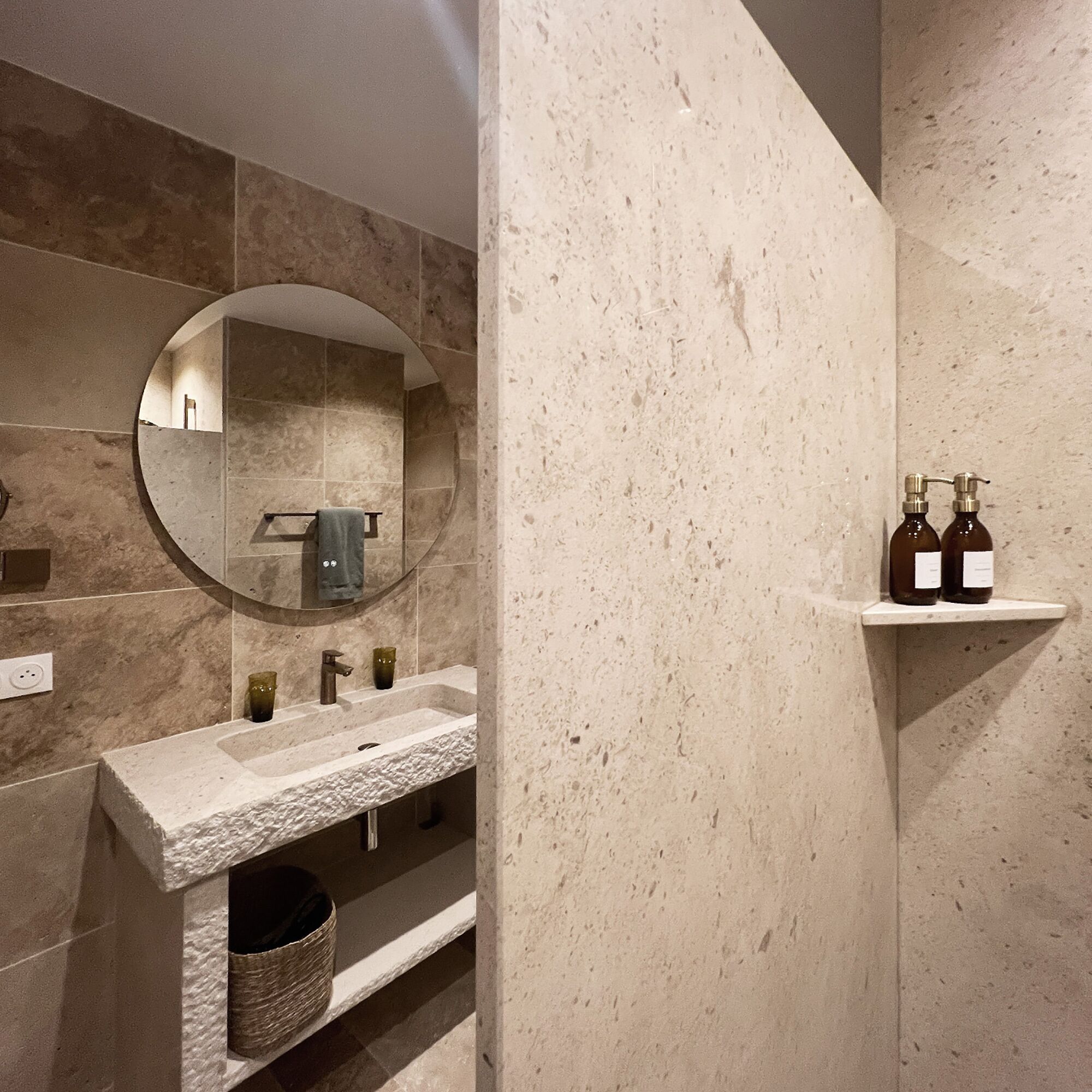 Une salle de bain avec un miroir rond, un lavabo et une étagère où sont posées des bouteilles.
