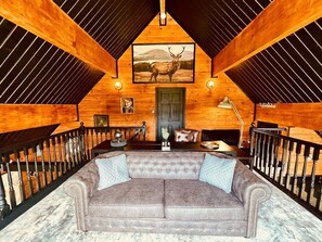 Interior de una cabaña de troncos con un sofá grande de color gris, paredes de madera, una imagen enmarco de un alce y una escalera con barandilla.