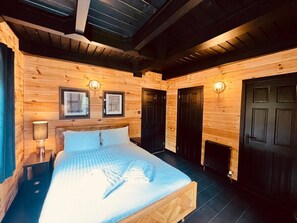 Un dormitori de cabana de fusta amb un llit, dues portes, una finestra i un lluminari fixat a la paret.