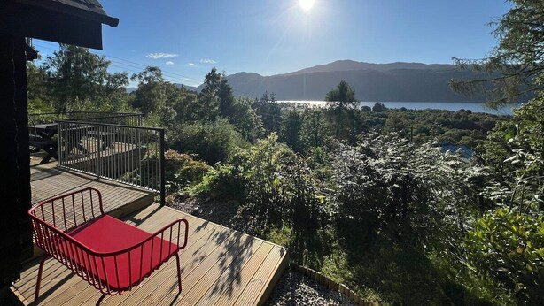 A wooden deck with a red chair, overlooking a mountainous landscape and a body of water.