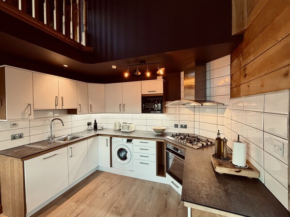 Une cuisine moderne avec des meubles blancs, un lave-linge encastré, une cuisinière à gaz et un escalier en bois.