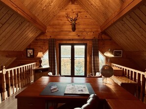 Un intérieur en bois avec un plafond haut, une tête de cerf accrochée au mur, une table avec une carte et un livre, et une vue sur une forêt par la porte.