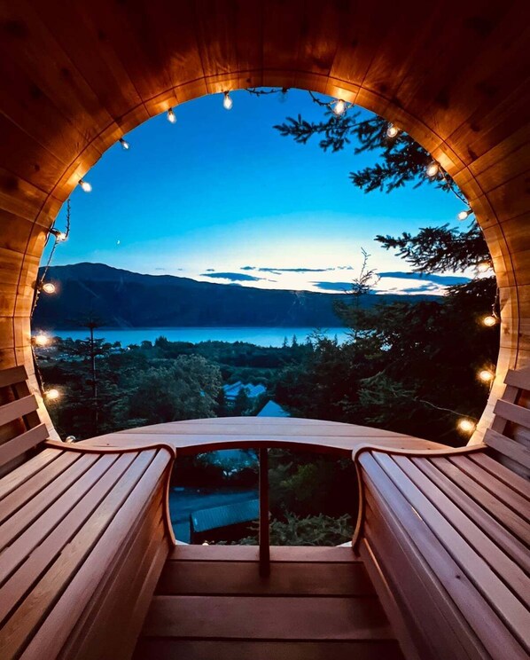 Una estructura de madera con techo curvo y un banco en su interior, con vistas a un lago y montañas.