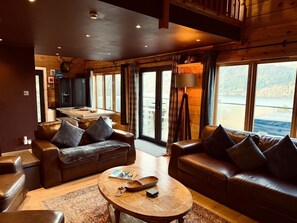 L’intérieur chaleureux d’un chalet avec des canapés en cuir, une table basse en bois et un coin cuisine visible à travers une grande fenêtre donnant sur un lac.