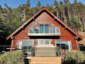 A two-story log cabin with a balcony, large windows, and a hot tub in front.