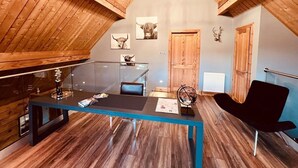 Un dormitorio estilo loft moderno con techo inclinado, vigas de madera, un escritorio de vidrio, una silla negra y un ventanal grande.