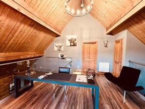 Une chambre de style loft moderne, avec un plafond en pente, des poutres en bois, un bureau en verre, une chaise noire et une grande fenêtre.