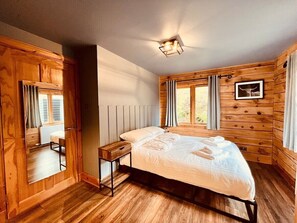 Una habitación de madera con cama, mesita de noche, espejo y ventana con cortinas.