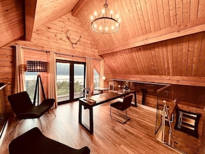 Un loft spacieux aux murs et au plancher en bois, un coin repas avec des chaises modernes, une grande fenêtre offrant une vue dégagée et un lustre.