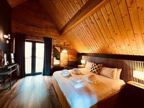 Une chambre accueillante avec un plafond en bois, un grand lit avec des serviettes soigneusement pliées, une table de chevet et une vue sur l’extérieur, accessible par une porte coulissante.