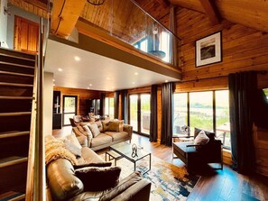 A spacious living area with a large sofa set, a coffee table, and a view of the outdoors through large windows. - Luxury Loch Ness lodge with hot tub & sauna (Inverness)