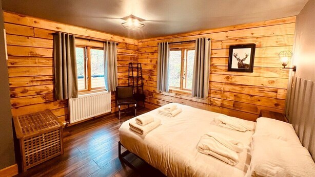Una habitación con paneles de madera, una cama, dos ventanas con cortinas, un radiador y un cuadro en la pared.