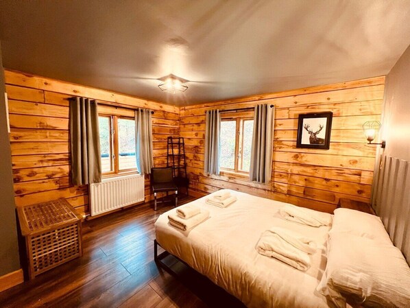 A wooden-paneled room with a bed, two windows with curtains, a radiator, and a framed picture on the wall.