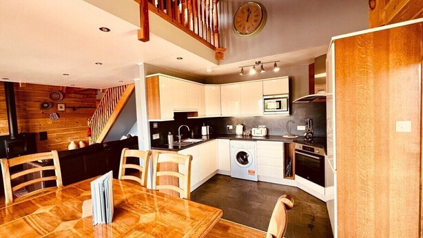 A compact kitchen with a dining area, wooden staircase, and a clock on the wall. - Group lodge with hot tub and fire pit (Inverness)