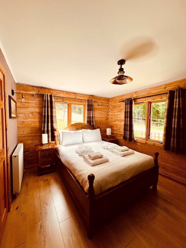 Un dormitorio con paneles de madera, una cama grande, dos mesitas de noche, un ventilador de techo y ventanas con cortinas.
