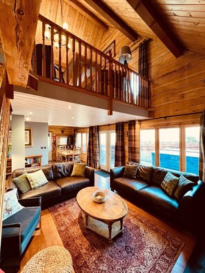A spacious living area with a high ceiling, wooden beams, a balcony, and large windows offering views.