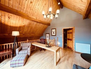 A cozy loft-style room with a high ceiling, wooden beams, a long wooden table, a plaid sofa, and a chandelier.