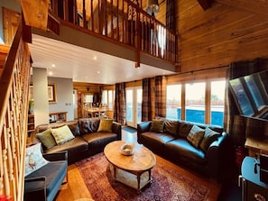 A spacious living room with a large sectional sofa, a coffee table, and a view of the outdoors through large windows. - Group lodge with hot tub and fire pit (Inverness)