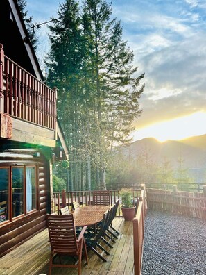A wooden deck with a table and chairs, overlooking a forest and mountains.