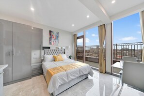A modern bedroom with a large bed, a balcony with a railing, and a city view. - Hotel Studio Apartment in East London (Ilford)