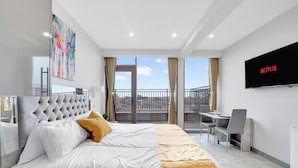 A modern hotel room with a large bed, a desk, a chair, a TV, and a balcony with a view. - Hotel Studio Apartment in East London (Ilford)