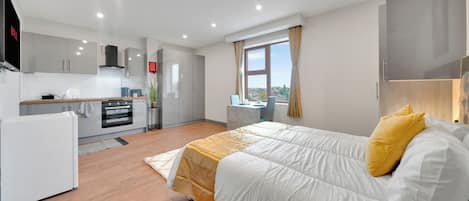 A modern hotel room with a large bed, a kitchenette, a dining area, and a view of the city.