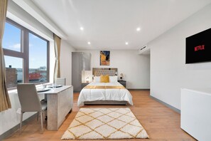A modern bedroom with a bed, desk, and chair, a large window, and a wall-mounted TV.