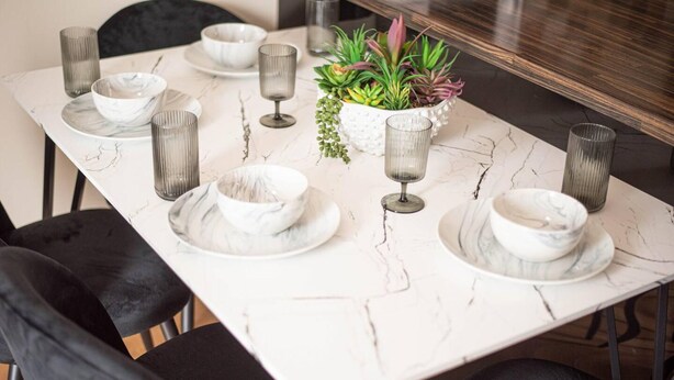 A dining table set with white plates, bowls, and glassware, accompanied by a potted plant and a marble-patterned tablecloth.