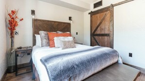 Quarto com cabeceira de madeira, uma cama grande com várias almofadas, uma mesa de cabeceira e um abajur fixado na parede.