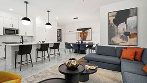 A modern kitchen with white cabinets, a dining area with black chairs, and a living room with a grey sofa and orange cushions.