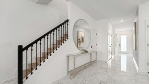 Un pasillo con piso de mármol, una escalera de madera con barandillas negras, una consola y un espejo grande.