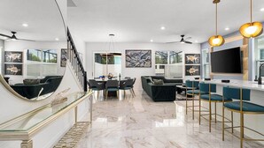 Una sala de estar moderna con piso de mármol, comedor con barandilla de vidrio, un sofá y una zona de bar con taburetes.