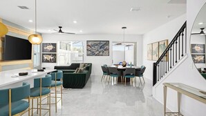 A modern living room with a sofa, a dining table with chairs, a kitchen area with a TV, and a staircase. - Villa Splendor | Brand NEW Villa w Designer Decor, Huge LR/DR, Pool - Near Parks (Orlando)