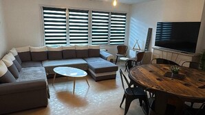 En stue med en hjørnesofa, et rundt sofabord, et spisebord med stole og et fladskærms-tv monteret på væggen.