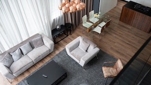 Uma sala de estar moderna com um sofá cinzento, um sofá cinzento menor, uma mesa de centro de vidro e uma poltrona de couro marrom. Há um rack de TV preto e um tapete cinza.