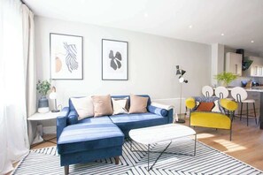 Ein modernes Wohnzimmer mit einem blauen Sofa, einem gelben Sessel und einer Küchenzeile im Hintergrund.